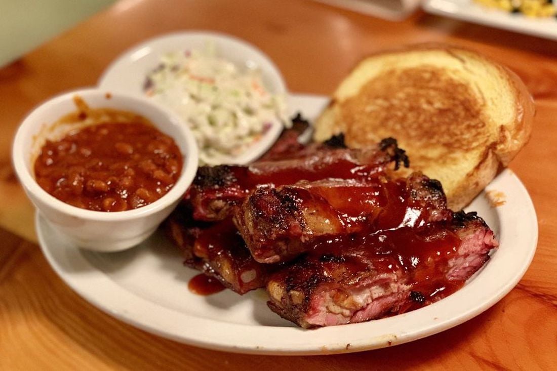 A plate with barbecued ribs covered in sauce, a slice of toasted bread, a bowl of baked beans, and a bowl of coleslaw on a wooden table.