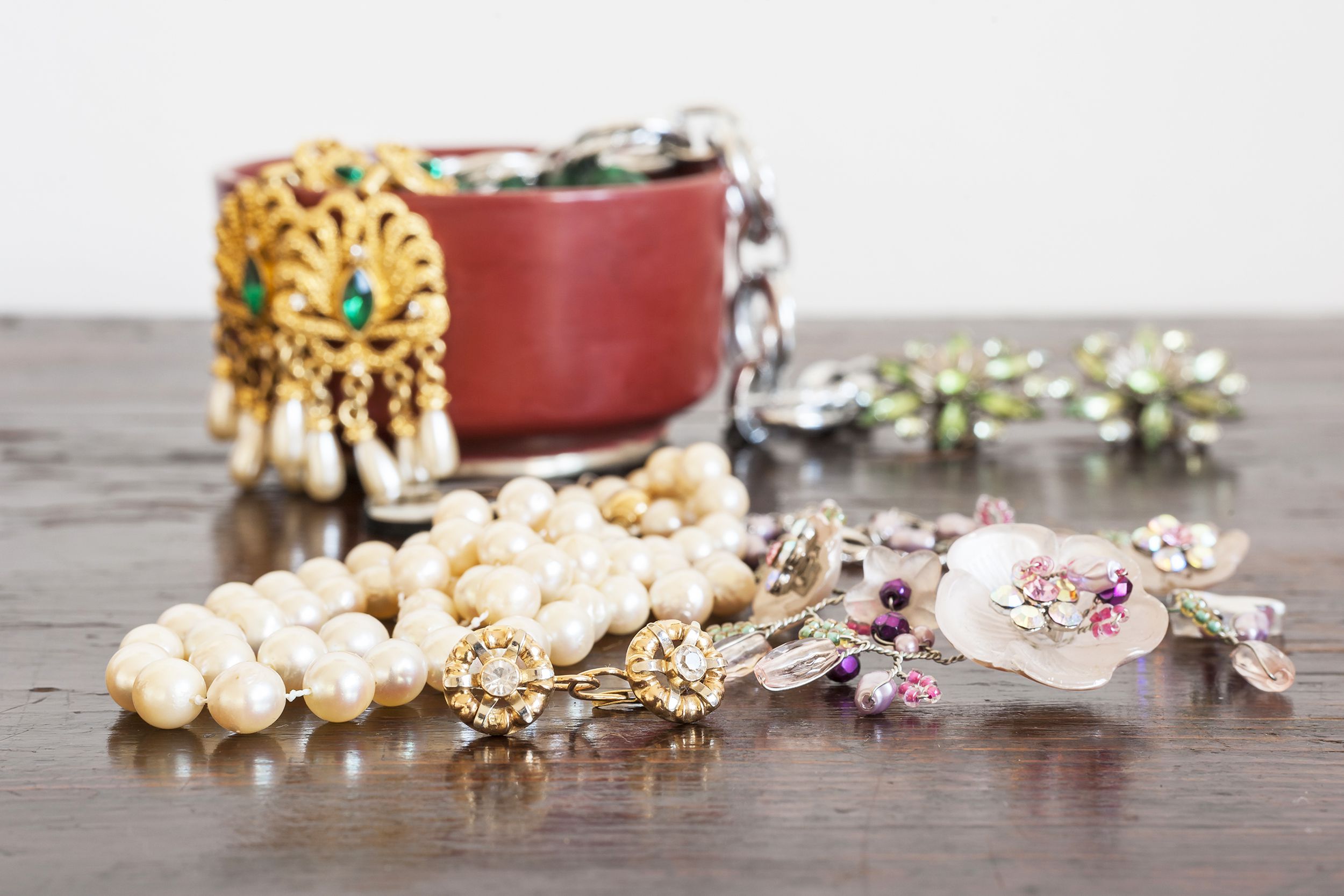 Closeup of costume jewelry, necklaces and earrings with a rust colored bowl filled with costume jewelry, pearls, diamonds, and flower designs, on a dark brown wooden table with a white wall in the background
