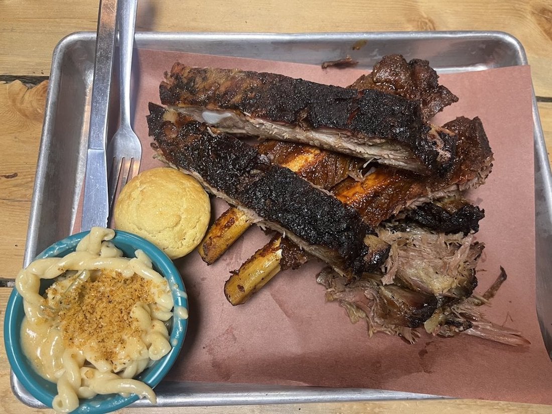 A tray with smoked ribs, a cornbread muffin, and a bowl of macaroni and cheese sits on brown paper on a wooden table. A fork and knife are placed to the side of the ribs.