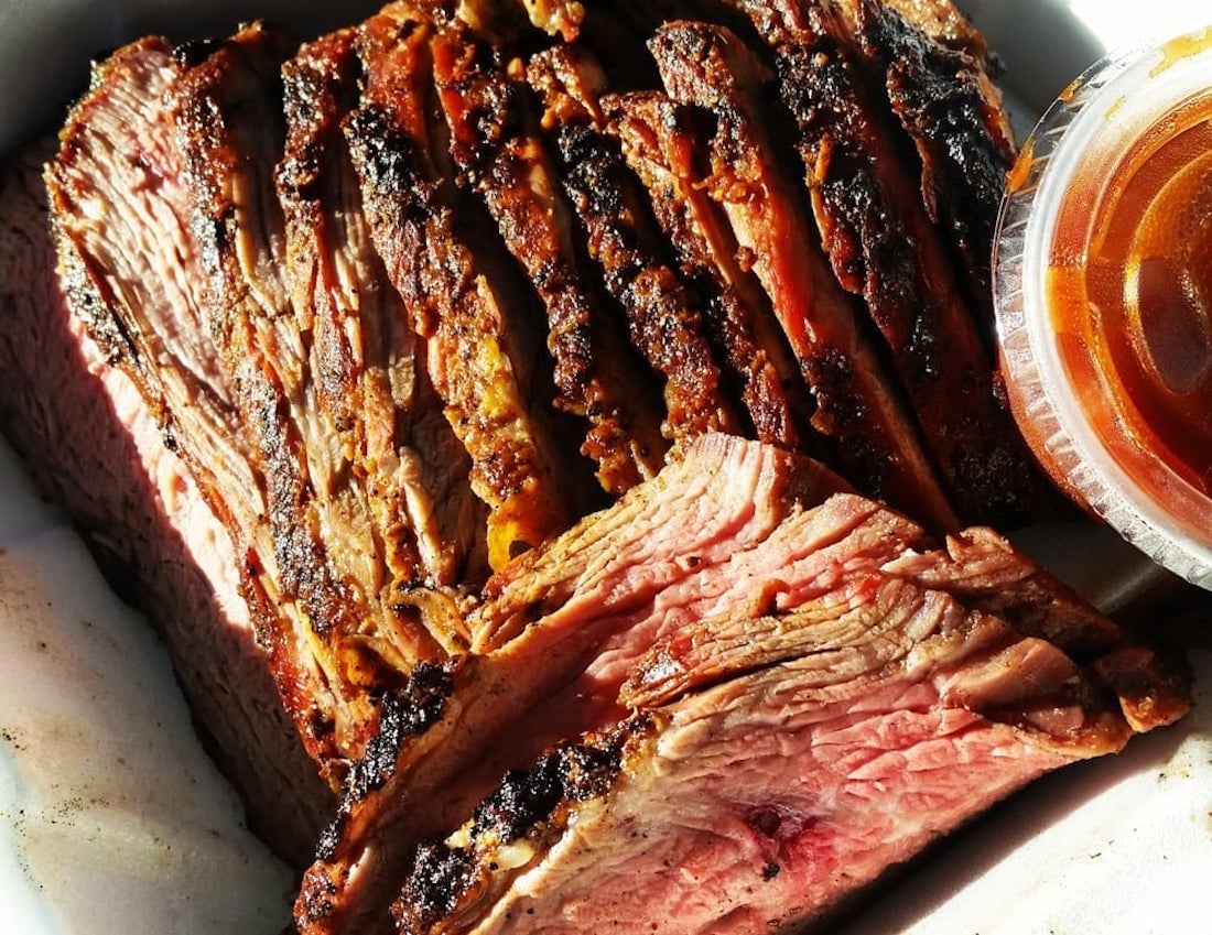 Sliced smoked brisket with a charred crust and juicy pink interior, arranged in a takeout container next to a cup of barbecue sauce.