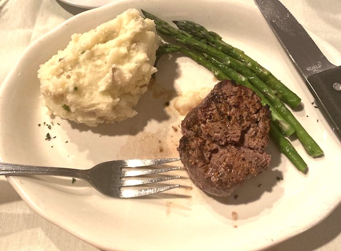 A plate with mashed potatoes, grilled asparagus, and a cooked steak filet. A fork rests on the plate near the potatoes, and a steak knife is on the edge of the plate. The food is partially eaten.