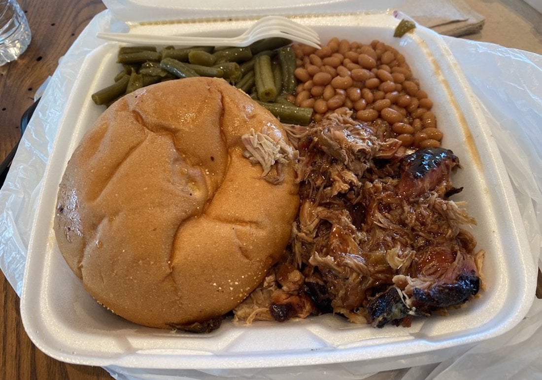 A takeout container with a pulled pork sandwich, green beans, baked beans, and a plastic fork on a napkin. The sandwich bun is partly covering the pulled pork, which is served next to the vegetables and beans.
