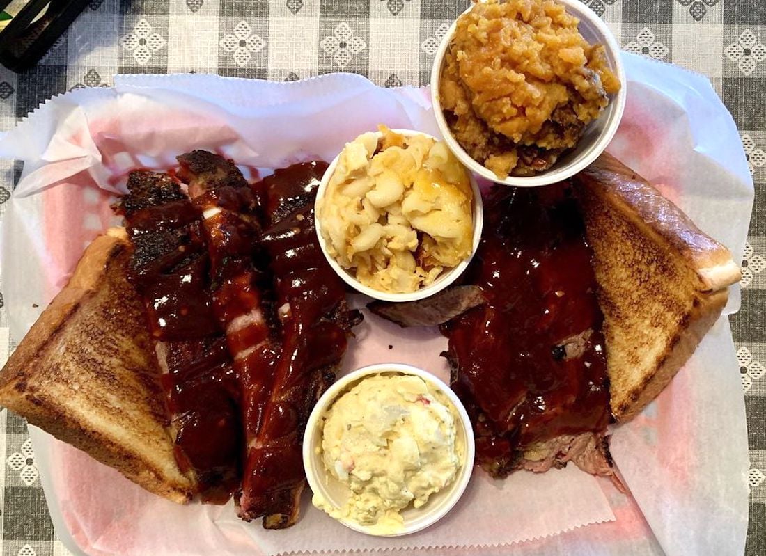 A tray with barbecued ribs and brisket covered in sauce, two slices of toasted bread, and three side dishes: macaroni and cheese, potato salad, and baked beans, on a white and gray patterned tablecloth.