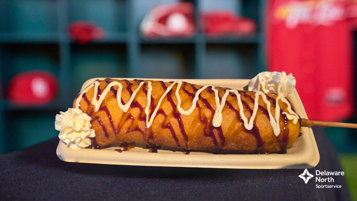 A deep-fried treat on a tray, drizzled with white and red sauces and topped with whipped cream swirls at both ends. A red jersey and cubby shelves are blurred in the background. Delaware North Sportservice logo is in the corner.