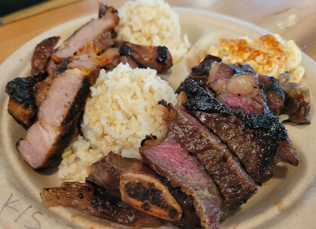 A plate of grilled steak slices, two scoops of rice, and a serving of macaroni salad, served on a round, disposable plate. The steak is seared with visible grill marks and pink centers.