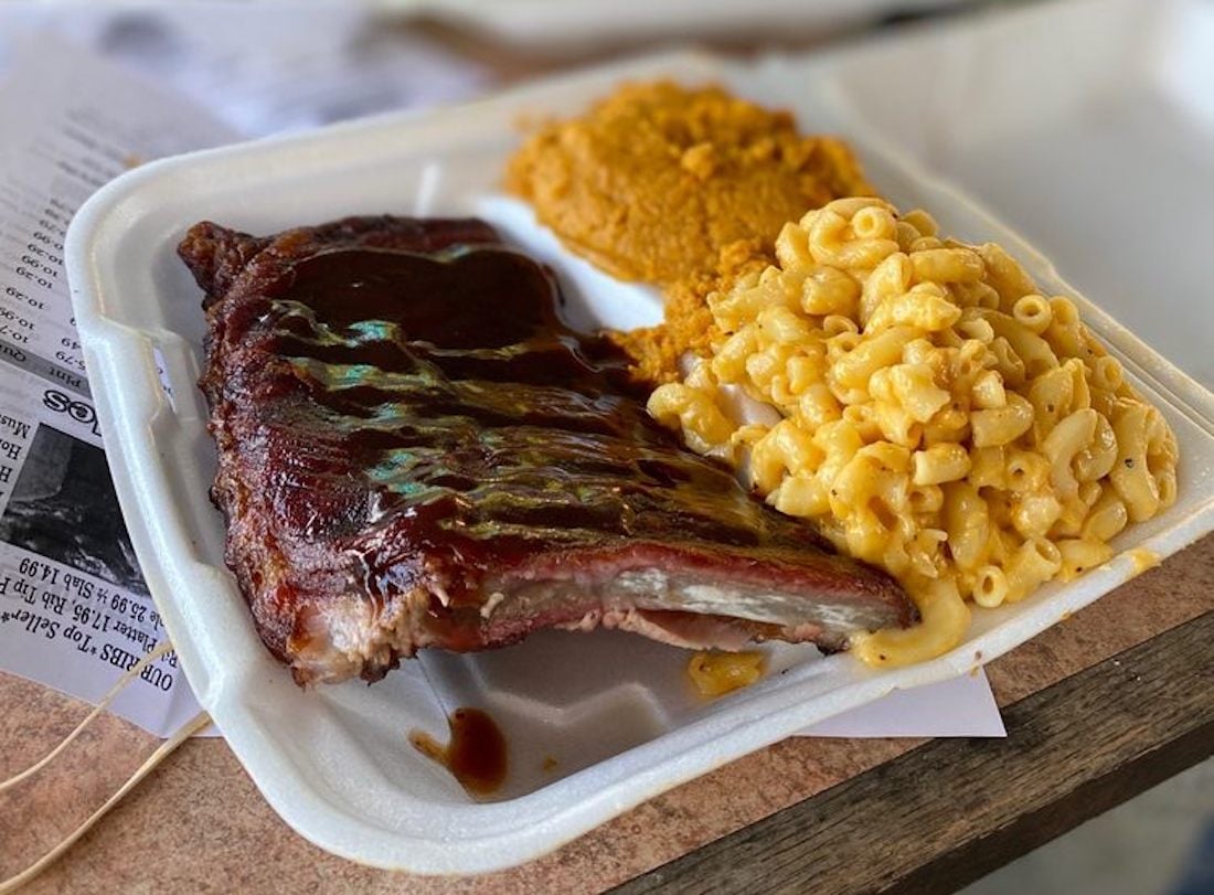 A takeout container with barbecue ribs covered in sauce, creamy macaroni and cheese, and a serving of mashed sweet potatoes, set on a table with some papers underneath.
