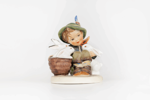A porcelain figurine of a smiling child in a green hat sitting beside a basket, surrounded by three white rabbits, on a round base against a white background.