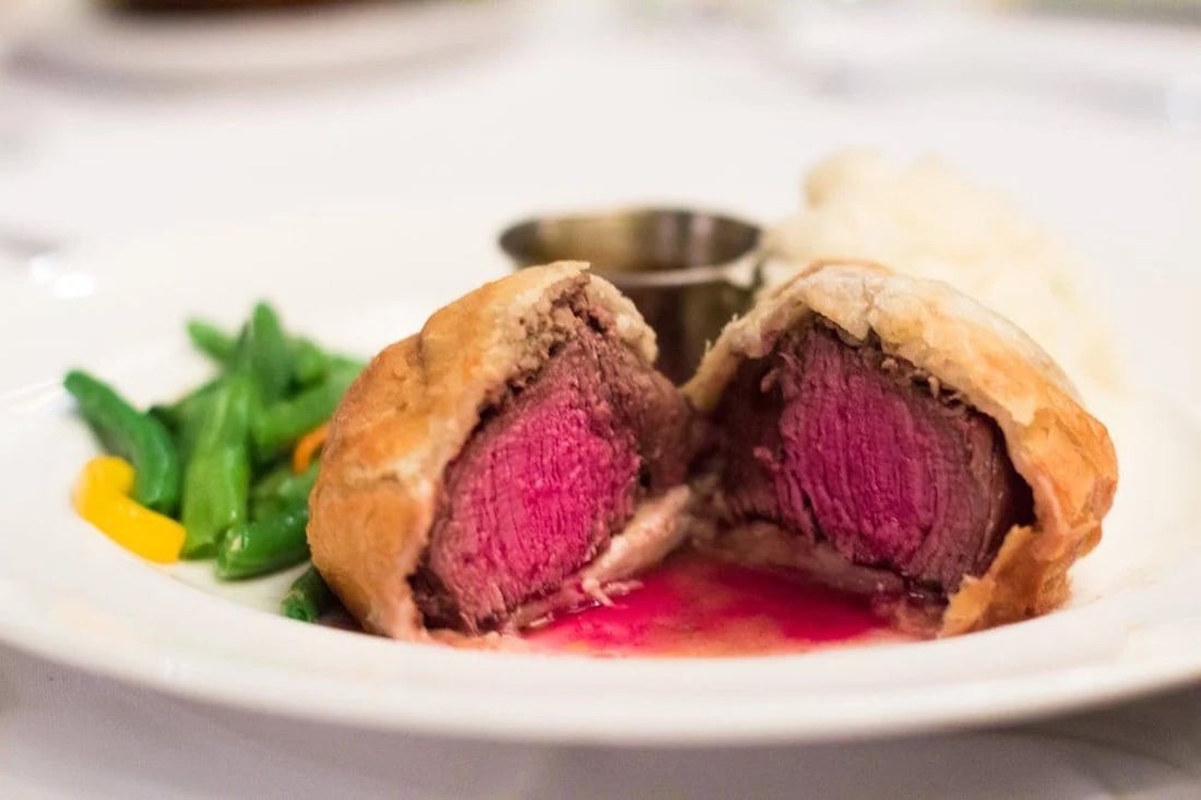 A sliced beef Wellington with medium-rare beef inside golden pastry, served on a white plate with green beans, yellow pepper strips, mashed potatoes, and a metal sauce cup in the background.