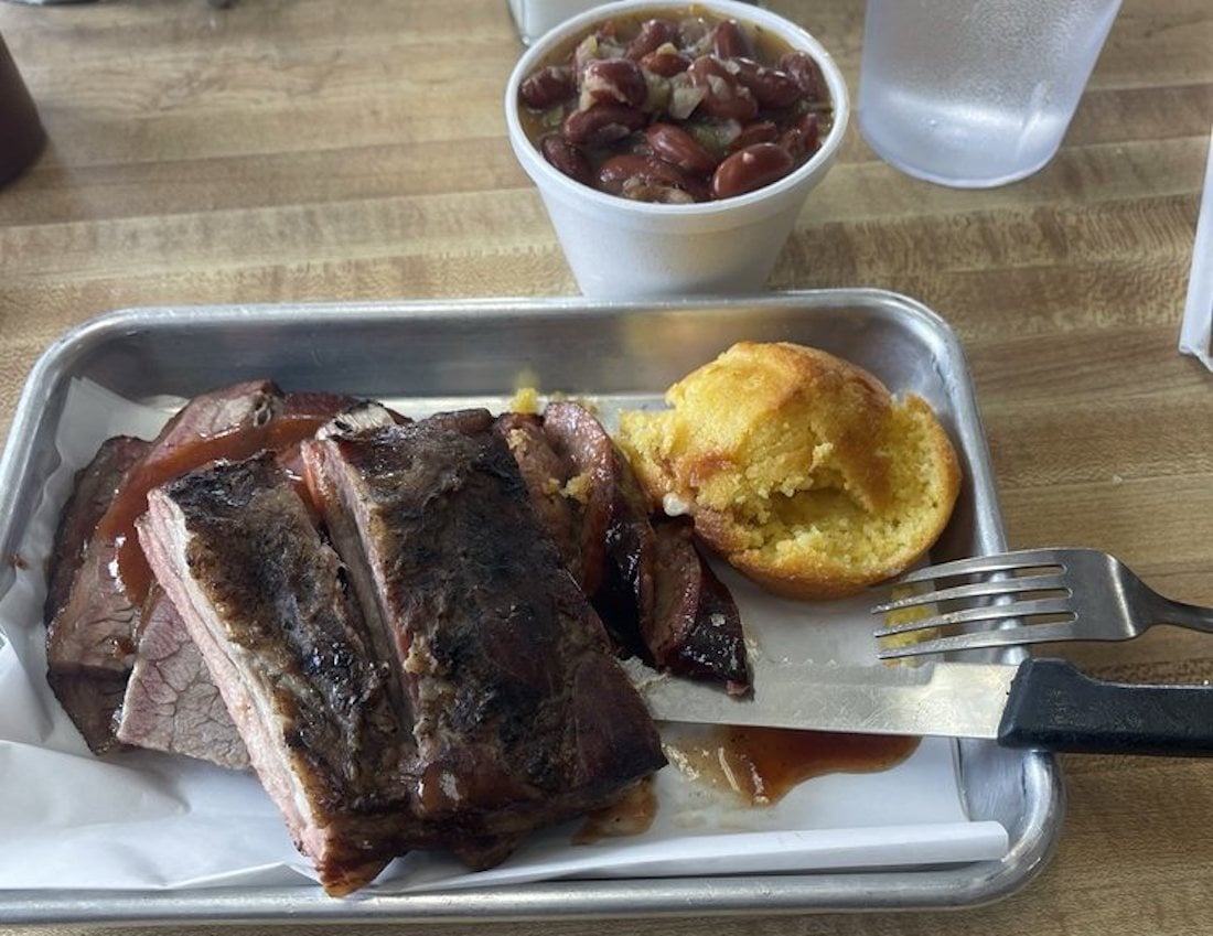 A metal tray with barbecue brisket slices, a piece of cornbread, and a fork. In the background is a cup of baked beans and a glass of ice water on a wooden table.