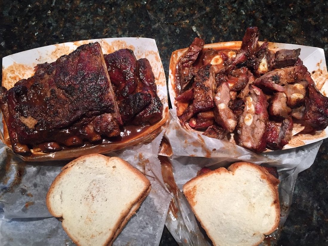 Two trays of barbecued ribs with sauce sit on a dark countertop, accompanied by two slices of white bread for each tray. One tray has whole rib slabs, the other has chopped ribs.
