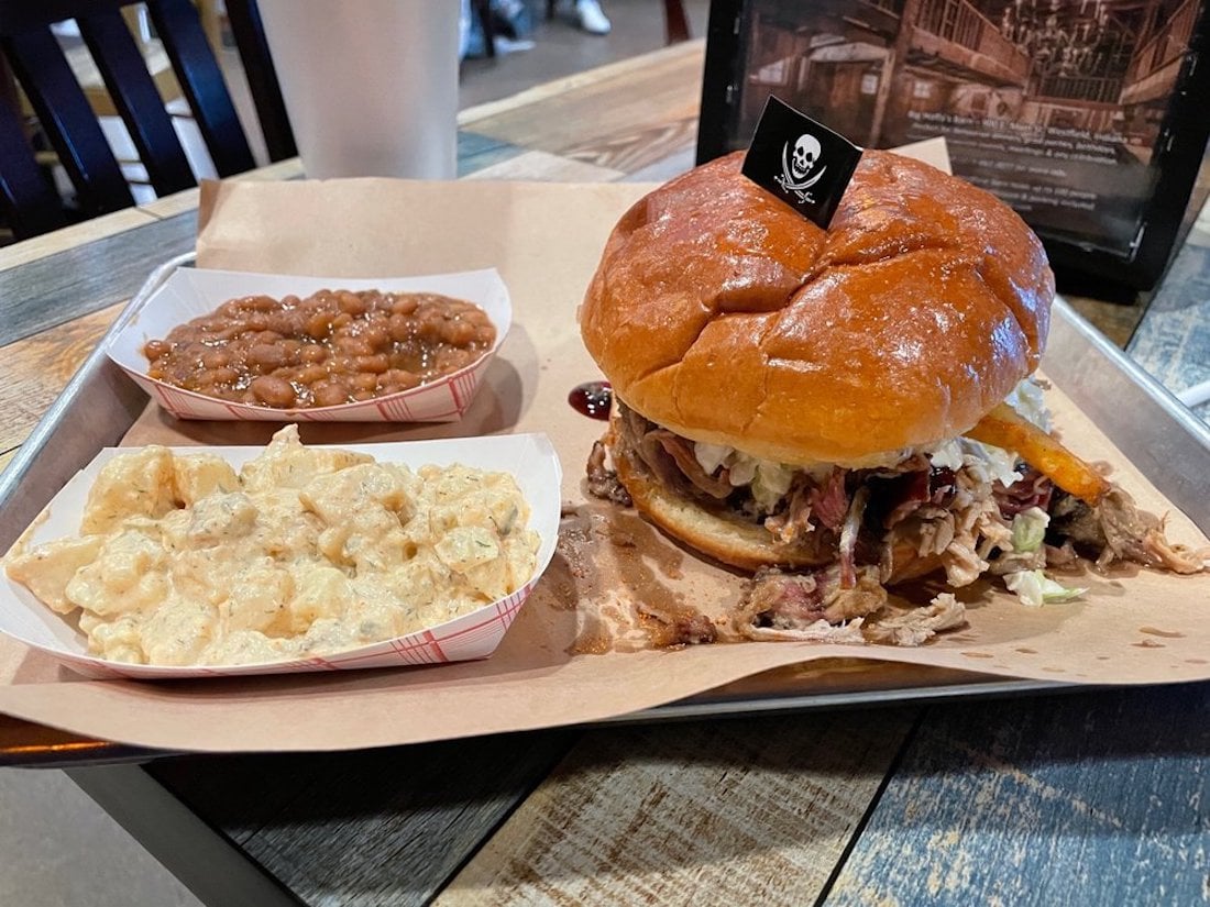 A tray with a pulled pork sandwich on a brioche bun, topped with a pirate flag, served alongside baked beans and potato salad in small paper trays. A drink cup is in the background on a rustic wooden table.