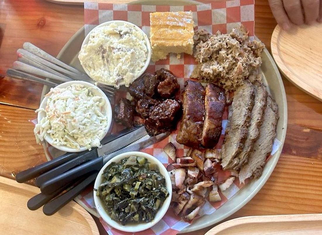 A large plate holds assorted barbecue meats, including brisket, ribs, pulled pork, and chopped chicken, with sides of collard greens, potato salad, coleslaw, cornbread, and several utensils on a wooden table.