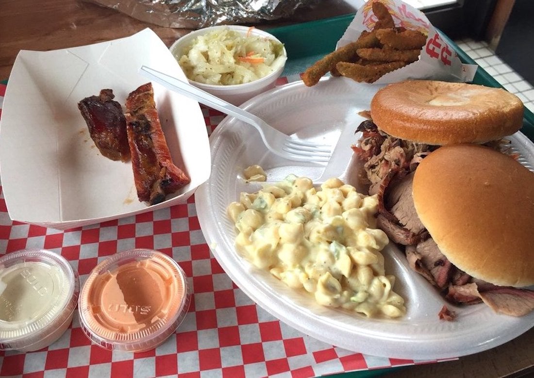 A plate of barbecue food including two sandwiches with pulled meat, macaroni salad, coleslaw, fried pickles, ribs, and two dipping sauces on a red checkered tray. A plastic fork is on the plate.