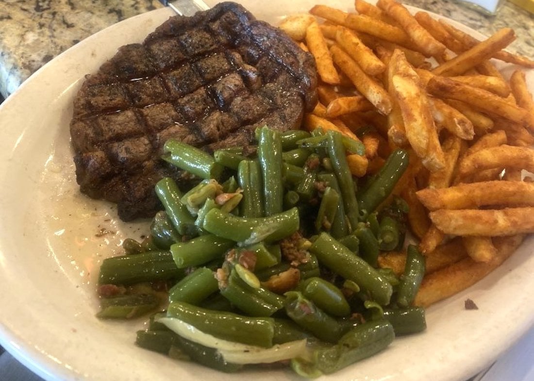 A plate of food featuring a grilled steak, golden French fries, and green beans mixed with bits of bacon and onions.
