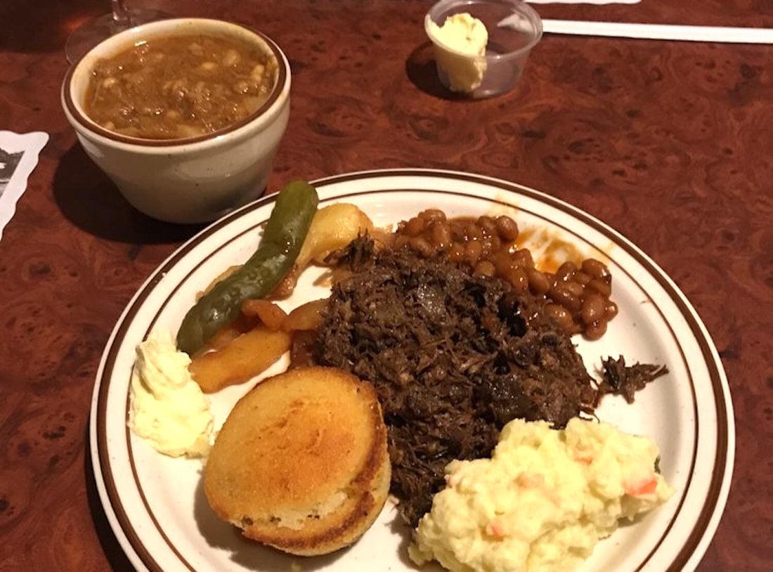 A plate with shredded beef, baked beans, potato salad, cornbread, cooked green peppers, and a pickle, next to a cup of brown stew and a small container of butter on a wooden table.