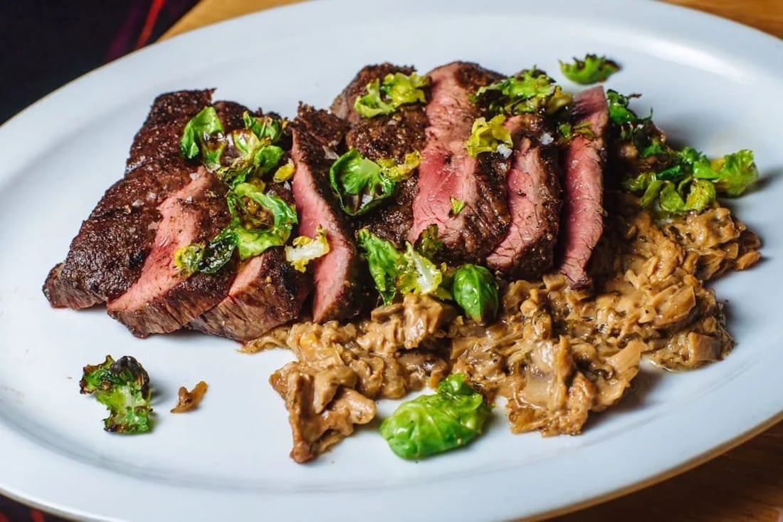 Sliced medium-rare steak served on a white plate with sautéed Brussels sprout leaves and a creamy mushroom sauce.