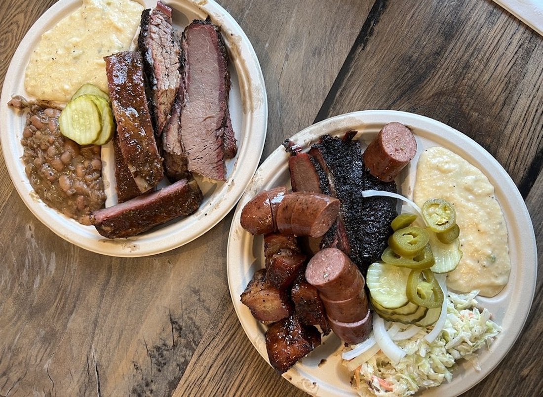Two plates of barbecue food on a wooden table. The plates have sliced brisket, sausage, ribs, pickles, beans, coleslaw, onions, jalapeños, and creamy yellow grits or corn pudding.