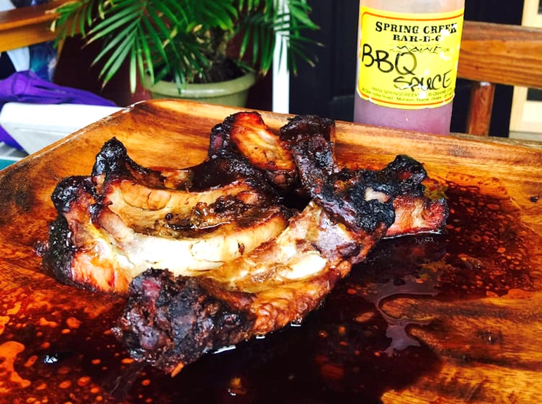 A wooden plate with juicy, charred barbecue ribs covered in sauce, next to a bottle labeled "Spring Creek Bar-B-Q BBQ Sauce." A plant and wooden chair are in the background.
