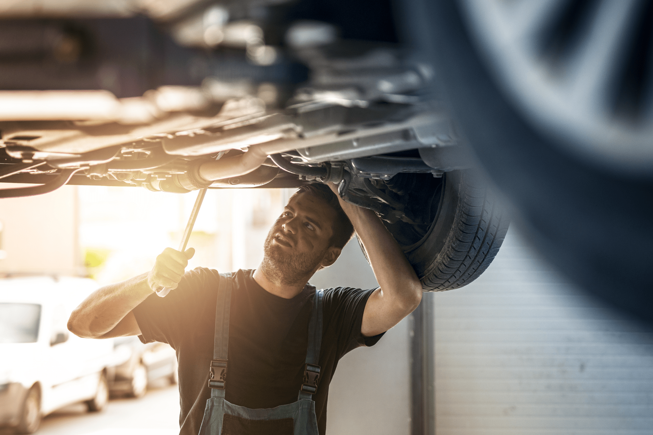 Male car mechanic repairing under elevated car with a tool, inside a repair garage with dramatic sunlight coming through from outside