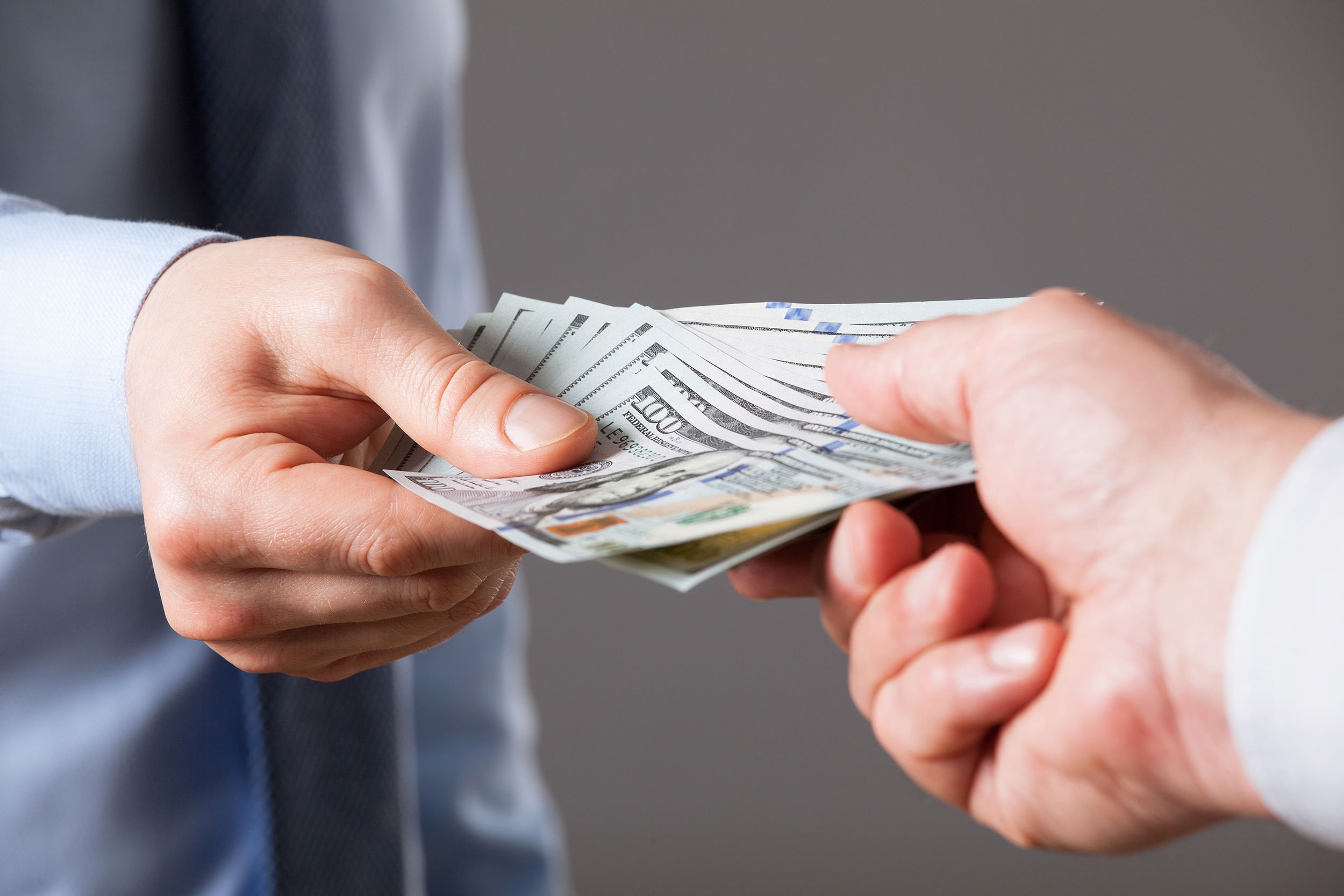 Closeup of Male hands giving numerous us $100 dollar bills to another male hand against neutral background