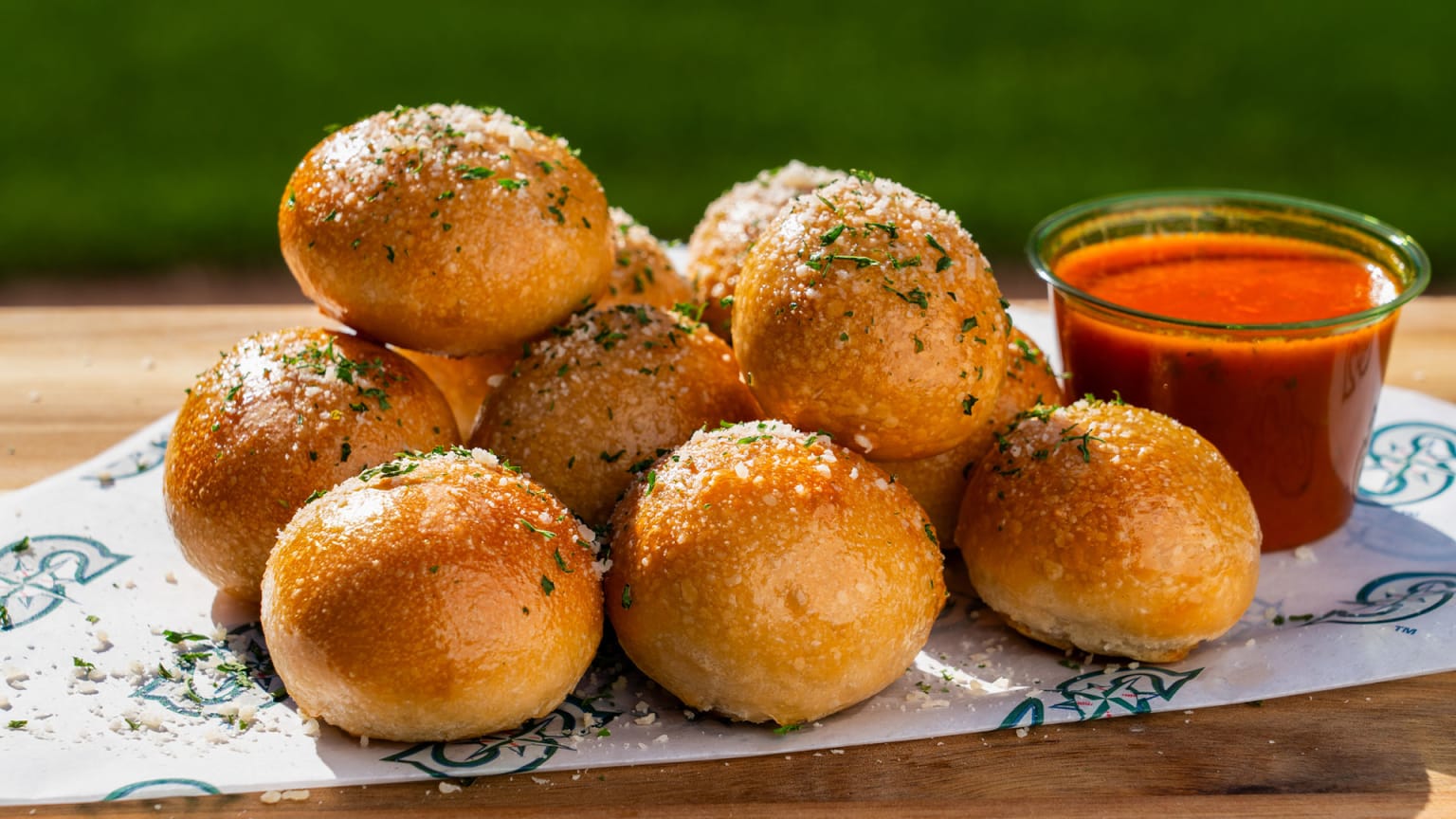 A pile of golden brown garlic knots topped with herbs and grated cheese sits on parchment paper next to a small glass cup filled with red marinara sauce. The background is blurred green grass.