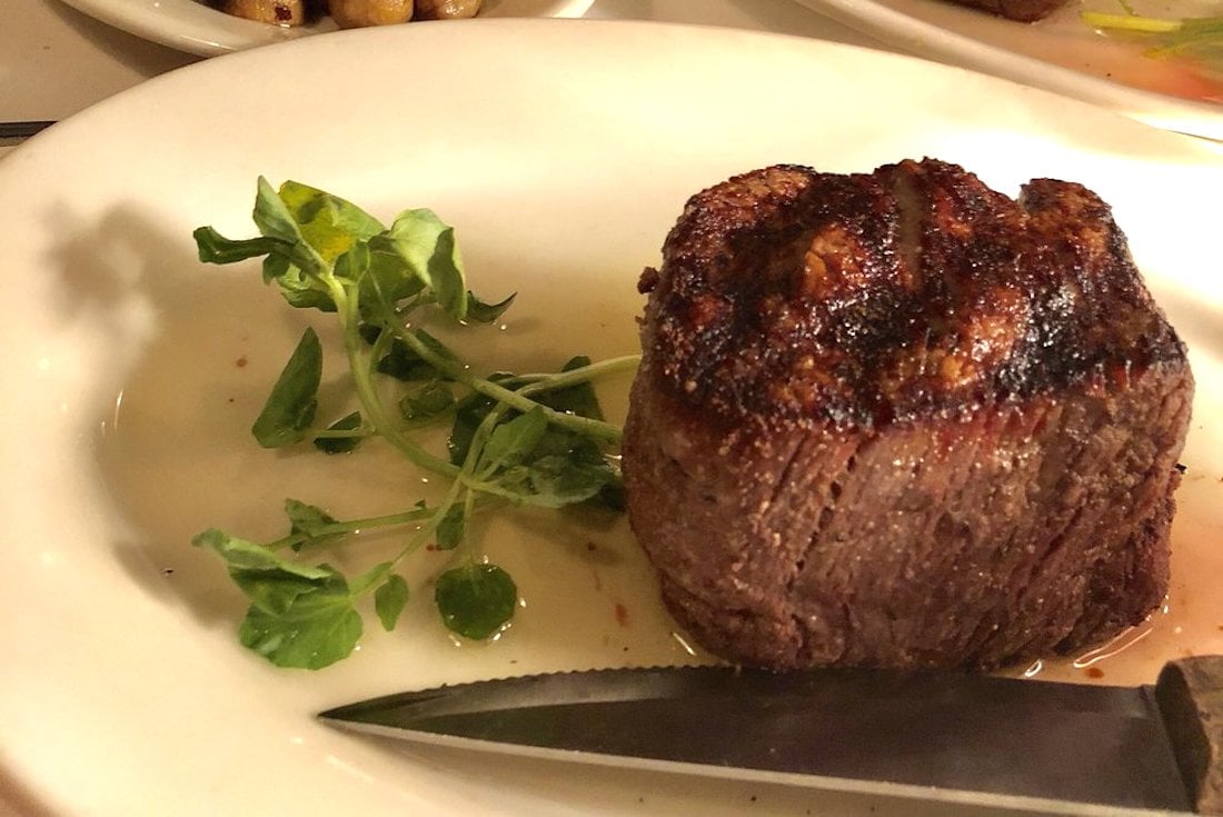 A cooked filet mignon steak sits on a white plate, garnished with a small sprig of greens and served with a steak knife.