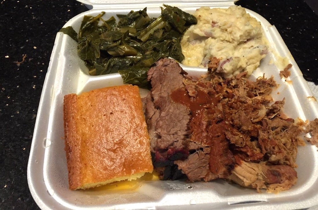 A takeout container with collard greens, mashed potatoes, cornbread, sliced brisket with barbecue sauce, and pulled pork on a black countertop.