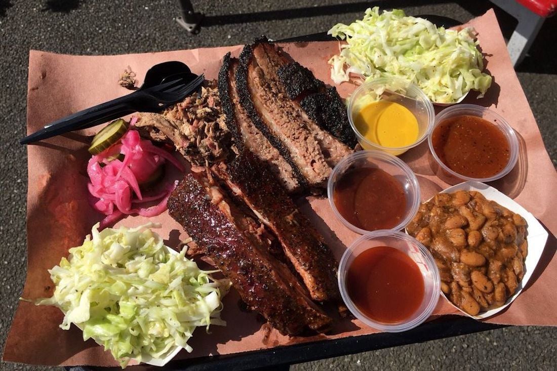 Barbecue platter with sliced brisket, ribs, pickled onions, pickles, coleslaw, baked beans, and four types of sauces in cups, all served on butcher paper with a black plastic fork and spoon.