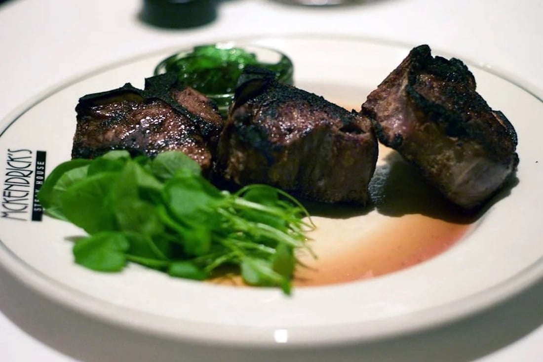 Three pieces of grilled steak sit on a plate next to a small bunch of fresh greens, with a small serving of sauce in the background. The plate is labeled “McKendrick’s Steak House.”