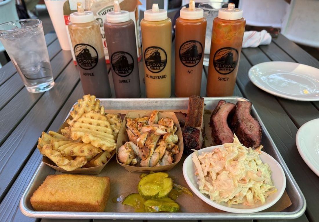 A tray of barbecue food with waffle fries, grilled chicken, ribs, coleslaw, pickles, and cornbread. Behind the tray are five squeeze bottles of different sauces. A glass of water and plate are on the table.