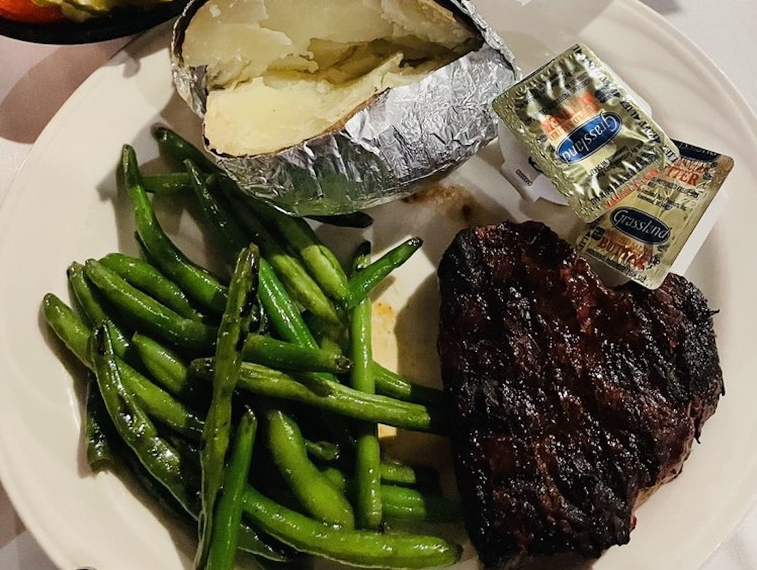 A plate with grilled steak, sautéed green beans, a baked potato wrapped in foil, and two packets of butter.