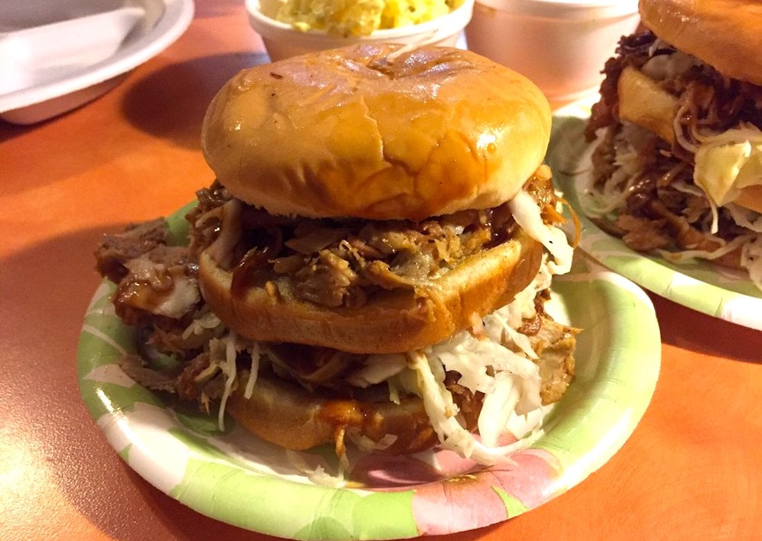 A pulled pork sandwich with coleslaw is stacked between a shiny toasted bun, served on a paper plate. Other food items and another sandwich are visible in the background on an orange table.