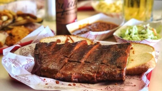 A tray of smoked ribs on sliced white bread, served with coleslaw, barbecue sauce, and fries in the background, along with a beverage and a barbecue sauce bottle.