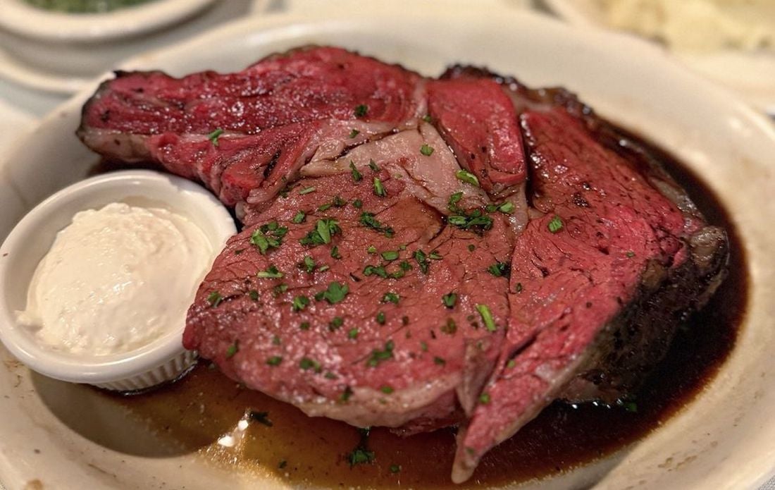 A thick slice of rare roast beef topped with chopped herbs, served in a shallow dish with dark juices and a small cup of creamy horseradish sauce on the side.