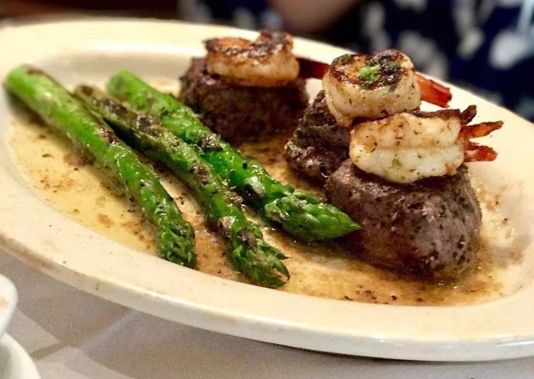 Grilled asparagus, two filet mignon medallions each topped with seared shrimp, served on a white plate with a light herb butter sauce.