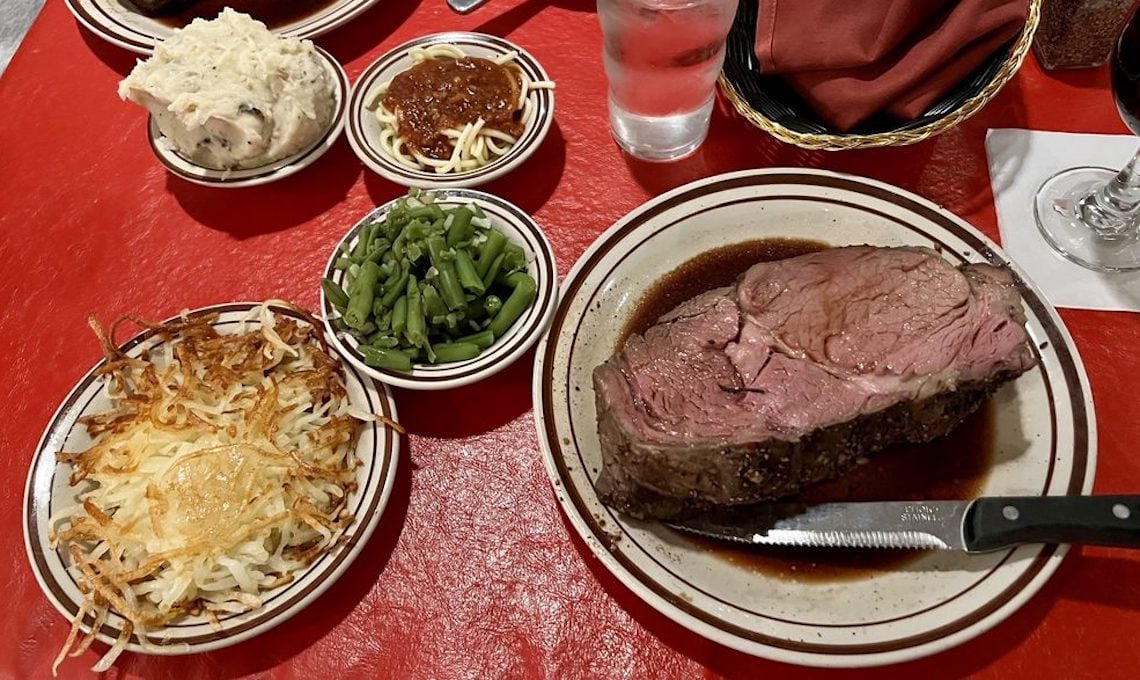 A plate of prime rib with au jus, surrounded by sides of hashbrowns, green beans, spaghetti with red sauce, and mashed potatoes. A glass of water and a glass of red wine are also on a red tablecloth.