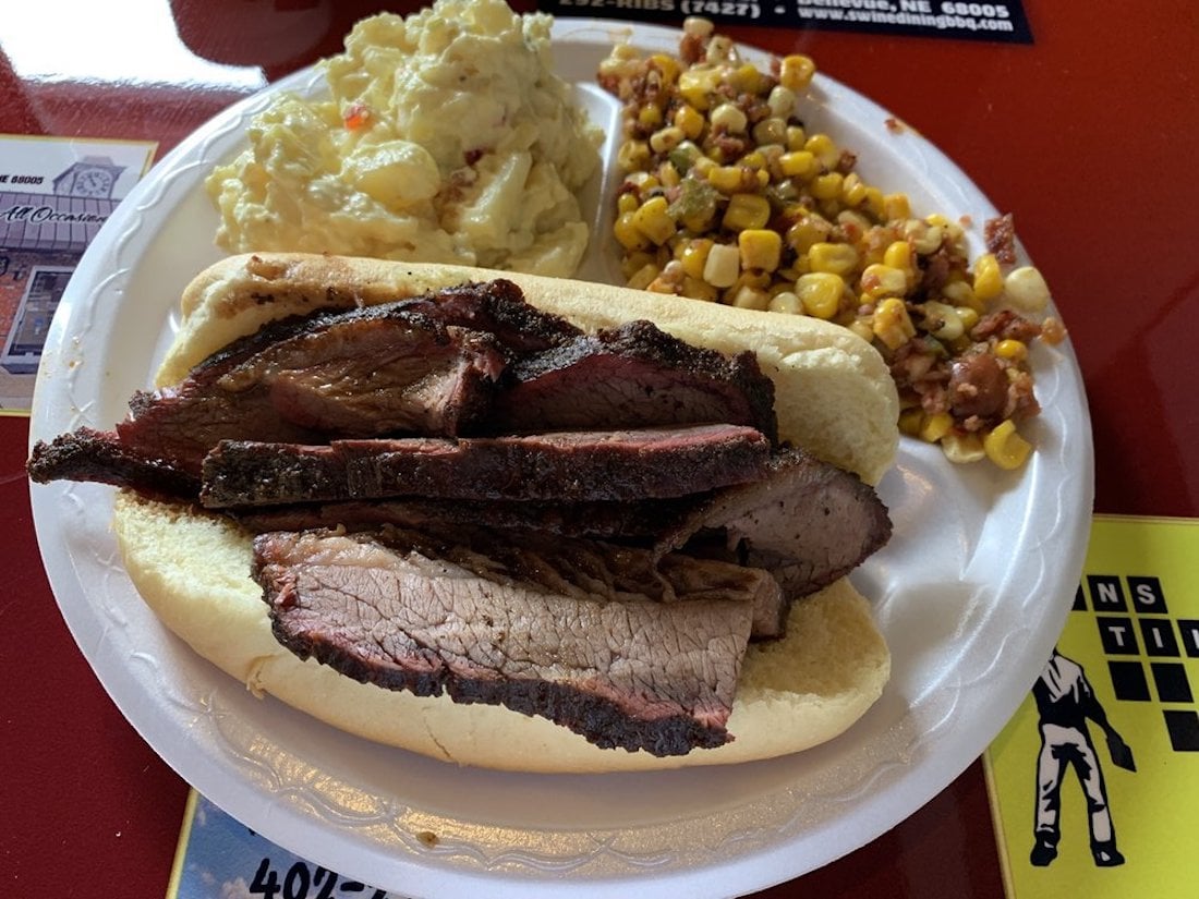 A white plate with a brisket sandwich on a bun, served with a side of potato salad and a corn and bean salad, on a red table.