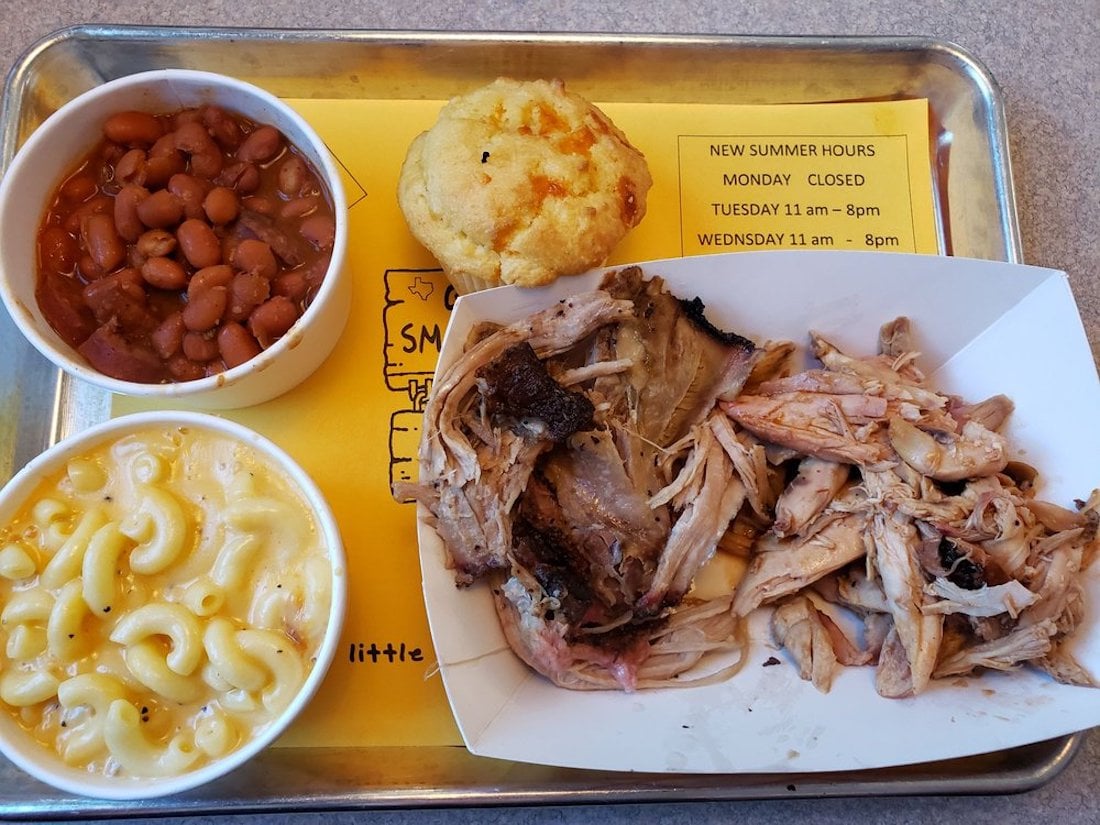 A tray with baked beans in a cup, a cornbread muffin, macaroni and cheese in a cup, and a paper tray of pulled pork barbecue, with a yellow sign showing restaurant hours underneath.