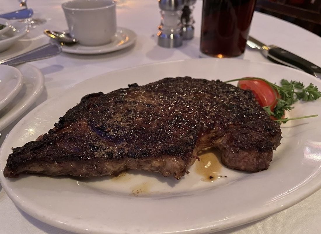 A cooked steak with a dark seared crust sits on a white plate, garnished with a sprig of parsley and a slice of tomato. The plate is set on a white tablecloth beside a cup and utensils.