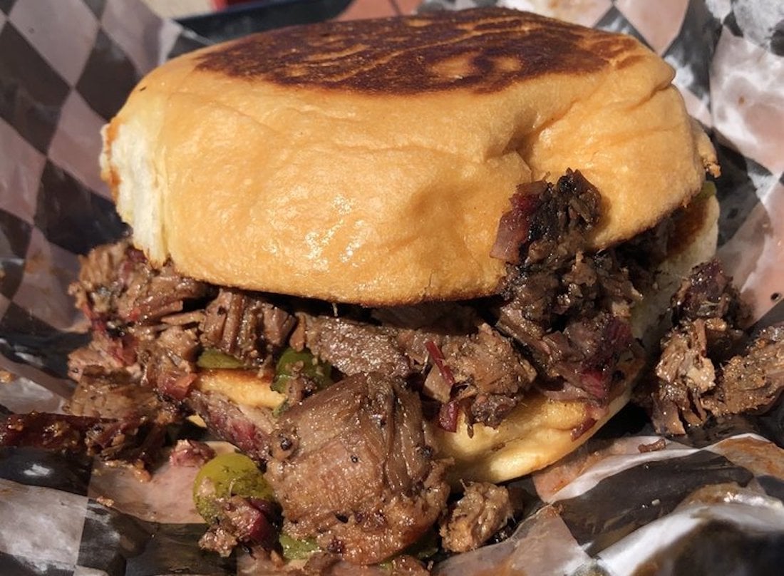 A close-up of a toasted sandwich bun filled with chunks of grilled or smoked beef, mixed with green peppers, on a checkered paper wrapper.