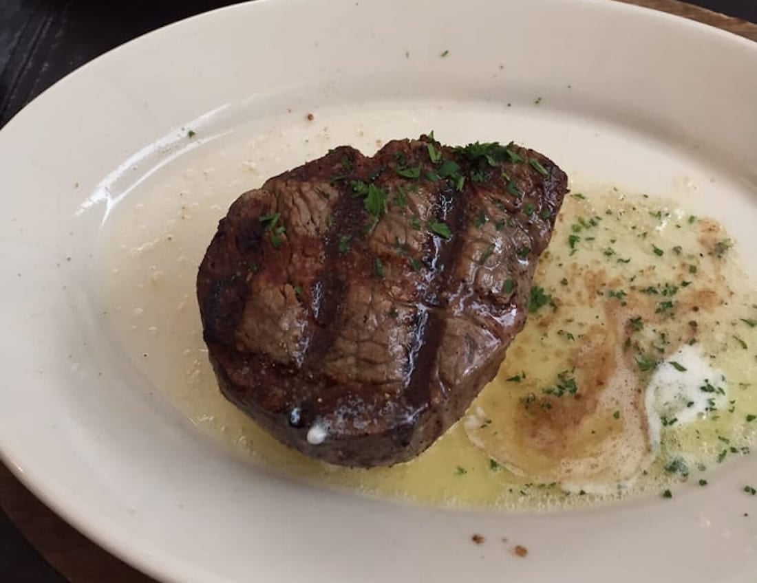 A grilled steak topped with chopped herbs sits on a white plate next to a pool of melted butter with herbs.