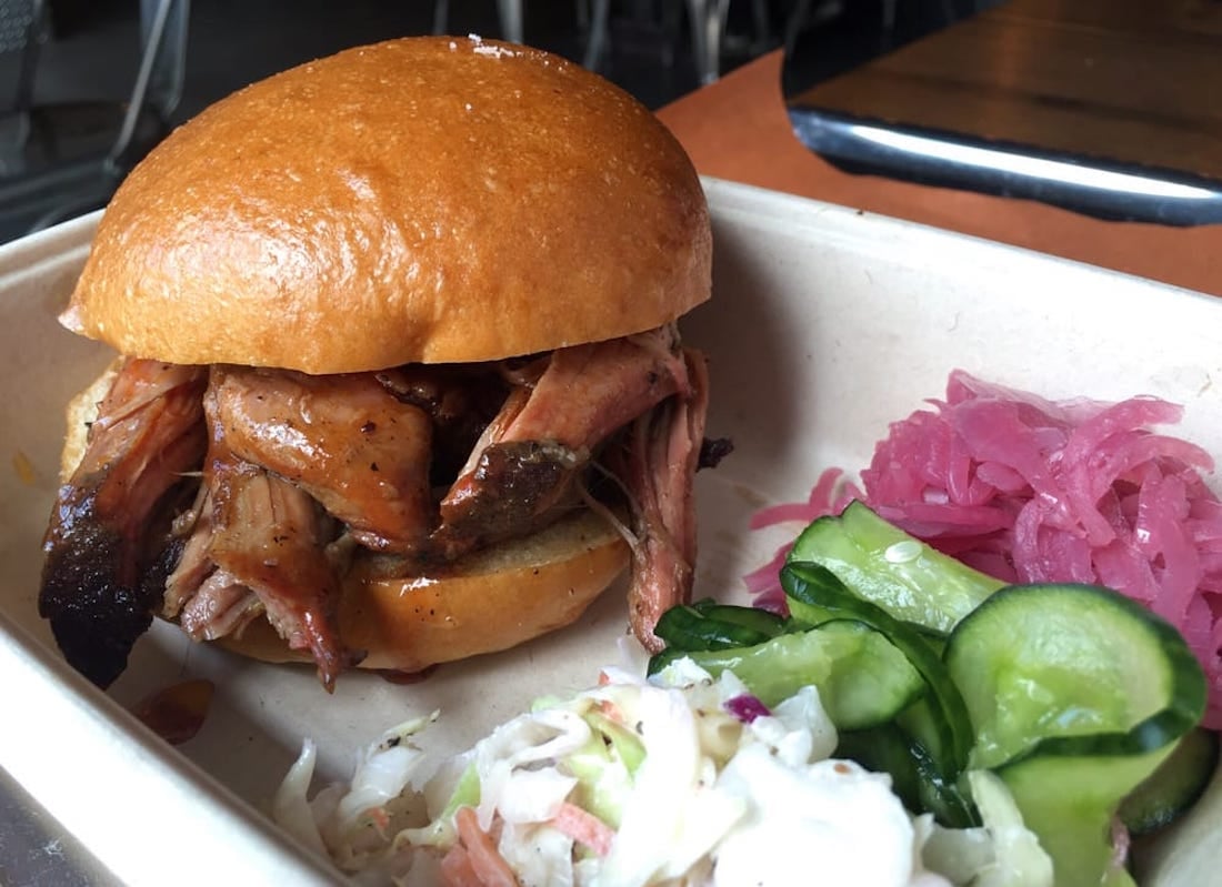 A pulled pork sandwich on a soft bun served with pickled red onions, sliced cucumbers, and creamy coleslaw on the side in a beige tray.