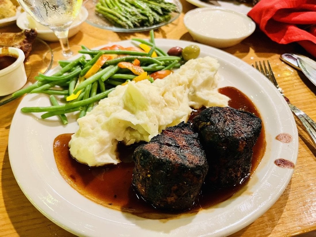 A dinner plate with two grilled steaks topped with brown sauce, mashed potatoes, and green beans with carrots. Additional dishes, asparagus, and a glass of water are visible in the background.
