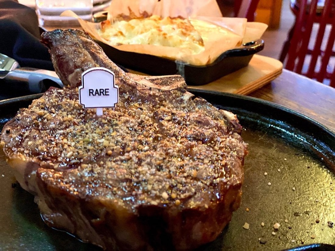 A cooked steak labeled "RARE" on a small marker sits on a black plate, seasoned with pepper. In the background, there is a dish of potatoes au gratin in a cast iron skillet.