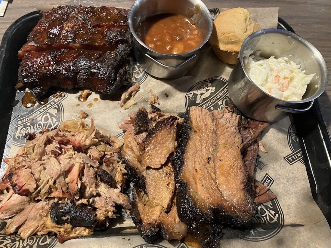 A barbecue platter with ribs, sliced brisket, pulled pork, baked beans in a cup, a biscuit, and a cup of coleslaw, all served on brown paper on a tray.