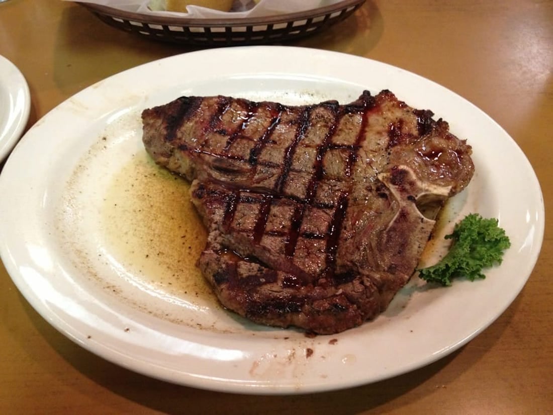 A cooked T-bone steak with grill marks sits on a white plate, with a small piece of parsley on the side and meat juices pooling around the steak.