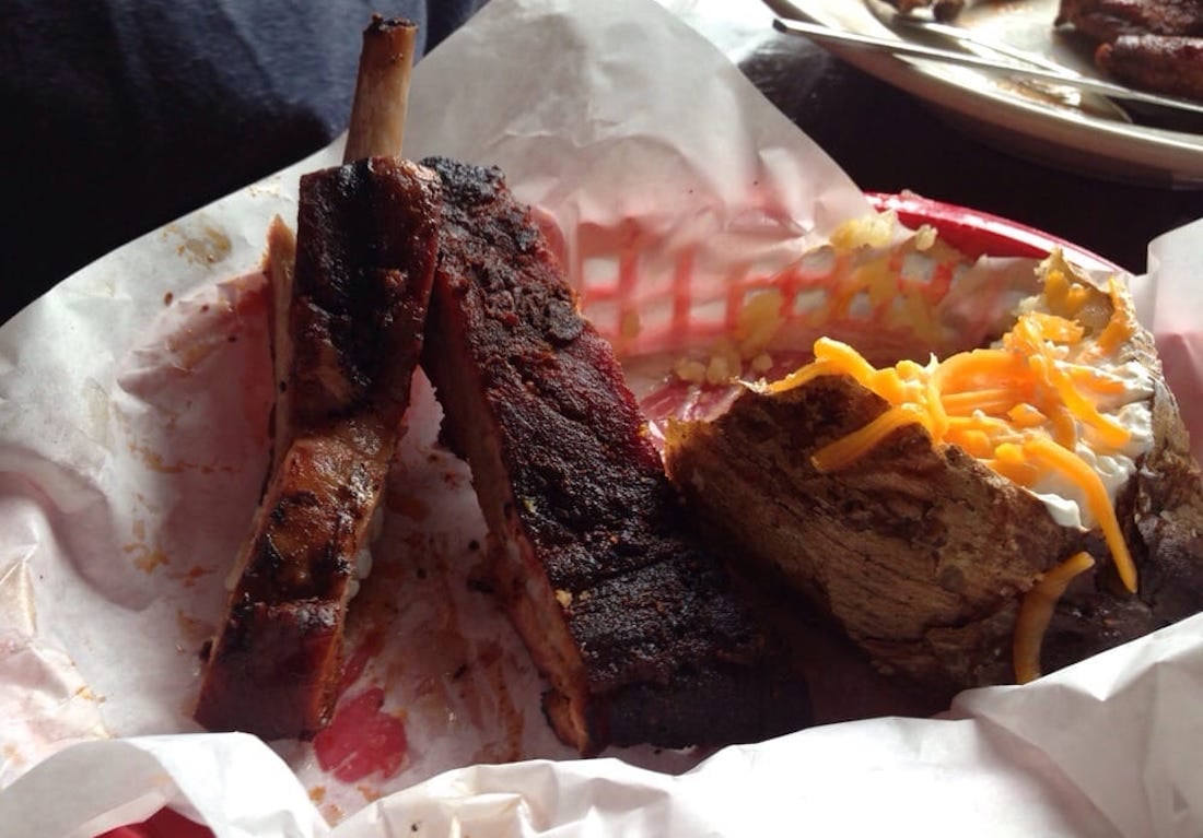 A red basket lined with paper holds two barbecued ribs and a baked potato topped with sour cream and shredded cheddar cheese.