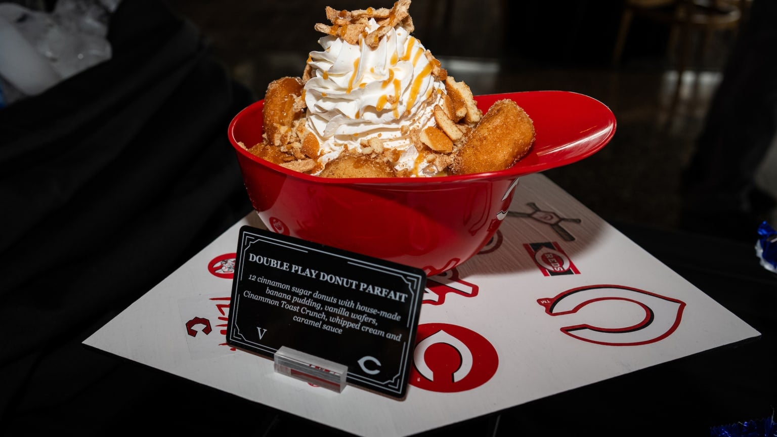 A red bowl filled with a donut parfait topped with whipped cream, caramel sauce, crushed nuts, and donut pieces. A sign in front reads “Double Play Donut Parfait.”