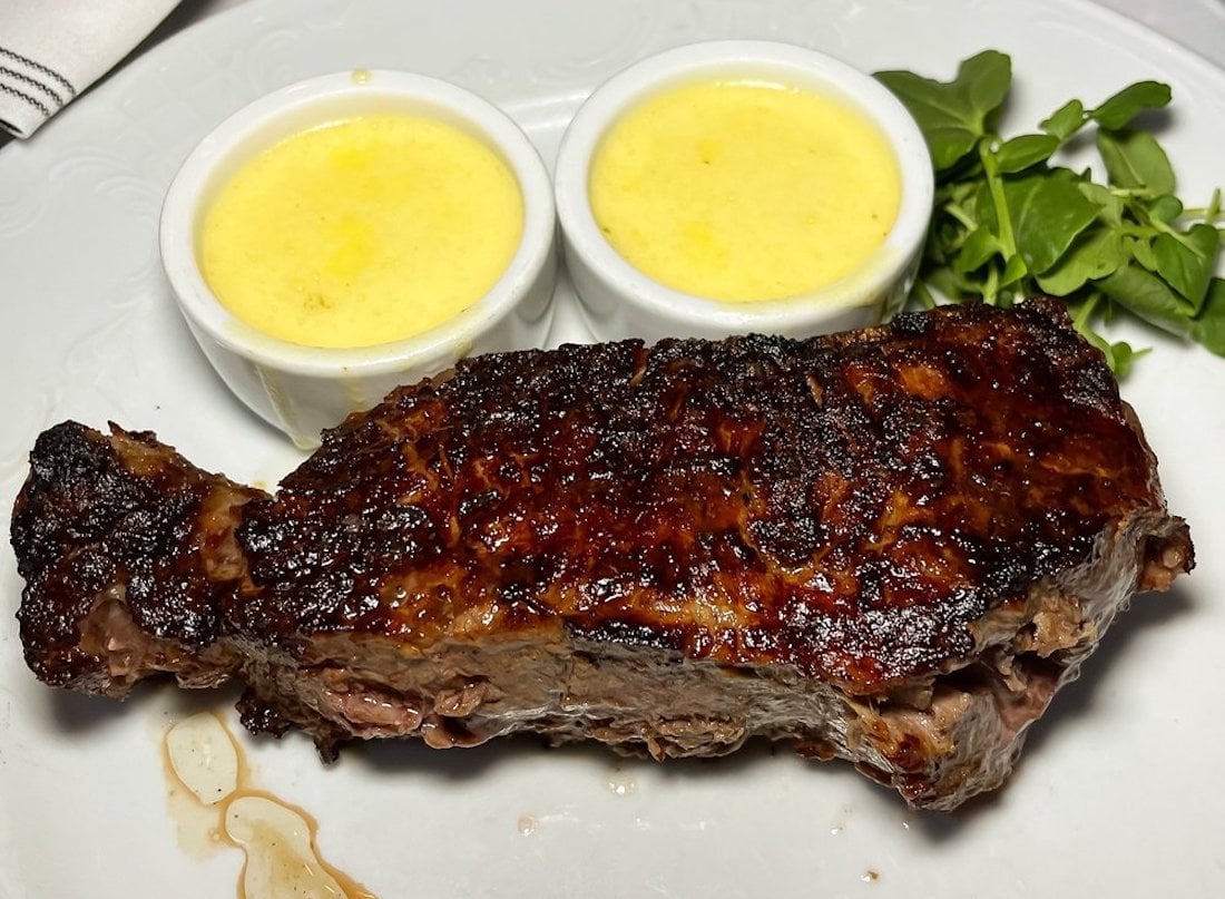 A grilled steak with a charred crust is served on a white plate alongside two small cups of yellow sauce and a small garnish of fresh greens.