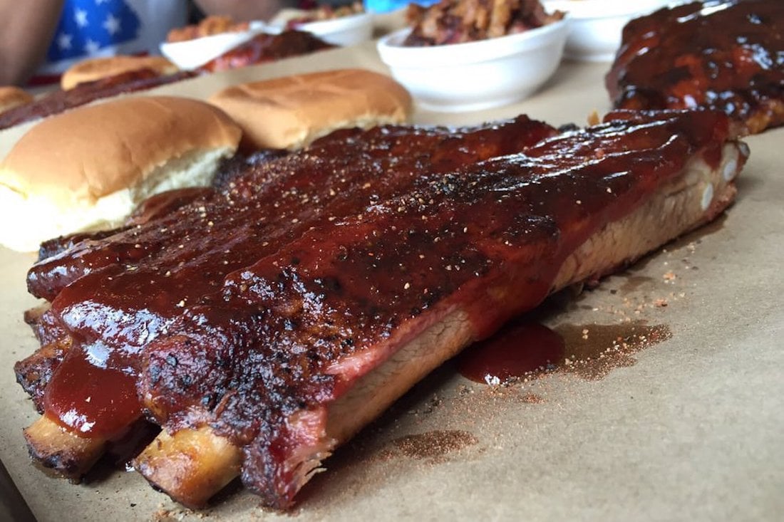 Barbecued pork ribs coated in sauce are placed on brown paper next to two sandwich buns, with bowls of food blurred in the background.